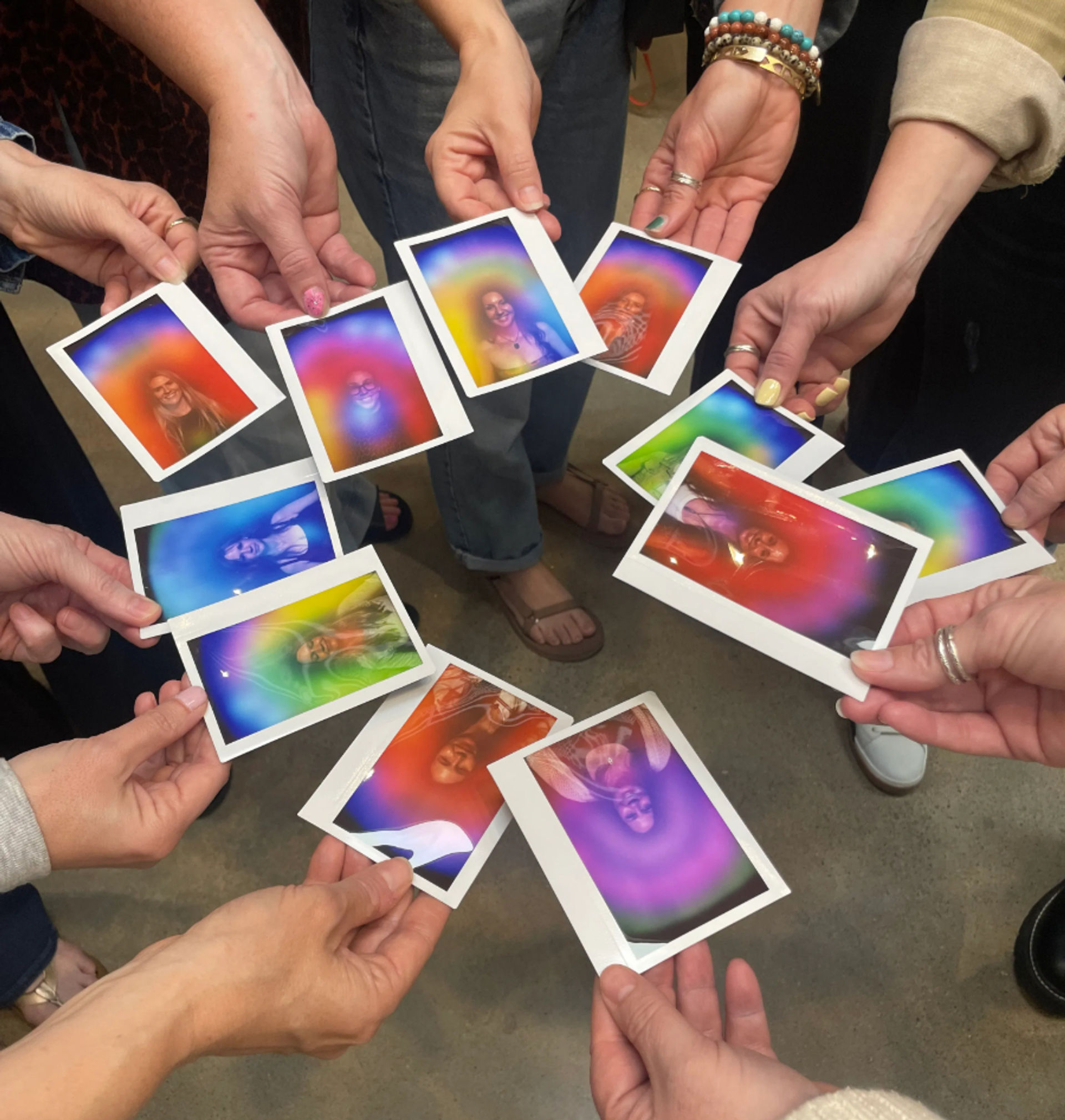 Overhead shot of a group holding instant Polaroid-style photos in a circle, each portrait surrounded by vibrant rainbow aura colors, hands and feet visible on a concrete floor — colorful group keepsake moment.
