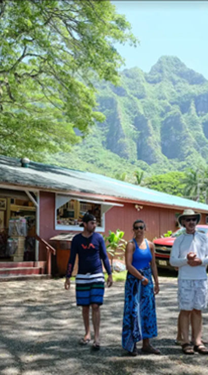 Three people strolling past a rustic red island market and parked pickup, framed by palm trees and dramatic green volcanic ridges under a bright blue sky — tropical island market scene.