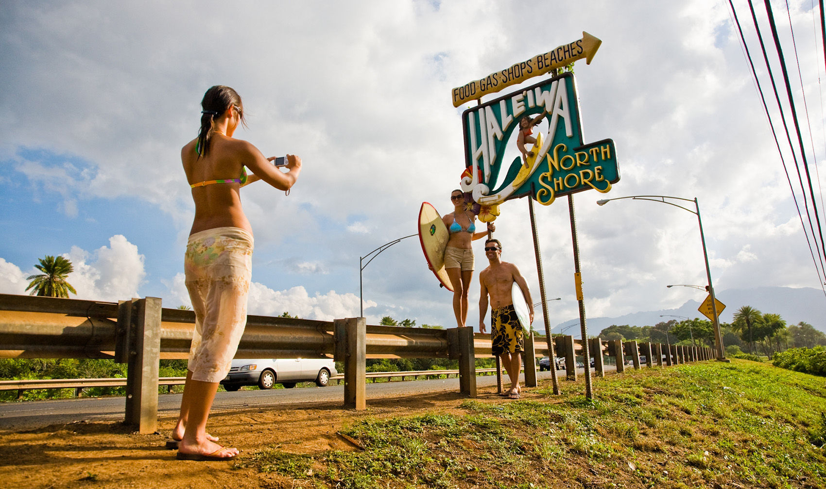 Woman photographing two surfers holding a surfboard beneath the colorful Haleiwa North Shore sign on Oahu’s North Shore, sunny roadside scene
