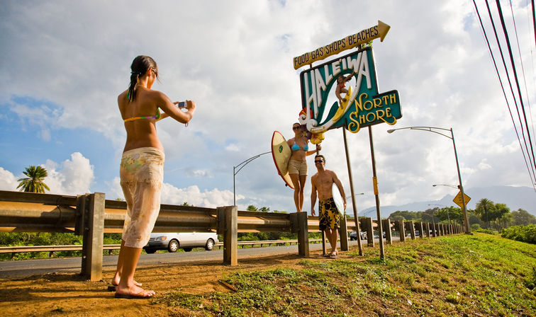 Woman photographing two surfers holding a surfboard beneath the colorful Haleiwa North Shore sign on Oahu’s North Shore, sunny roadside scene