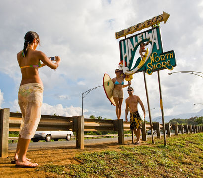 Woman photographing two surfers holding a surfboard beneath the colorful Haleiwa North Shore sign on Oahu’s North Shore, sunny roadside scene