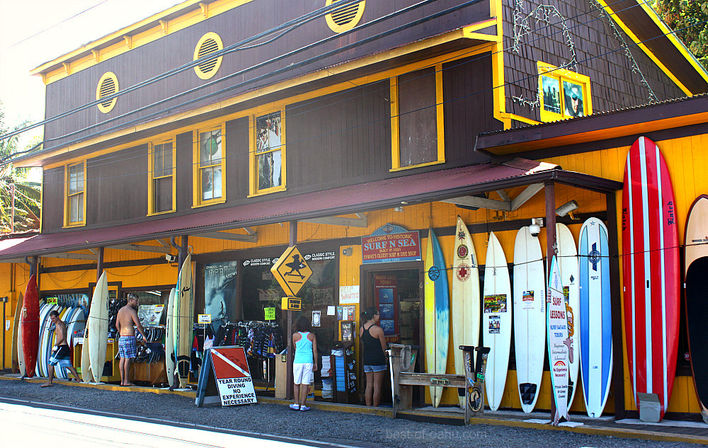 Bright coastal surf shop storefront with colorful standing surfboards, wetsuit and gear racks, and people browsing under a yellow-and-brown awning on a sunny street — beach town surf store scene.