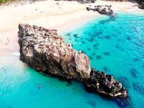Aerial view of a tropical sandy beach with vivid turquoise water, a large rocky outcrop and swimmers near the rocks