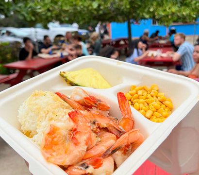Takeout tray with plump cooked shrimp, white rice, sweet corn and a pineapple wedge held over red picnic tables at a casual outdoor dining area