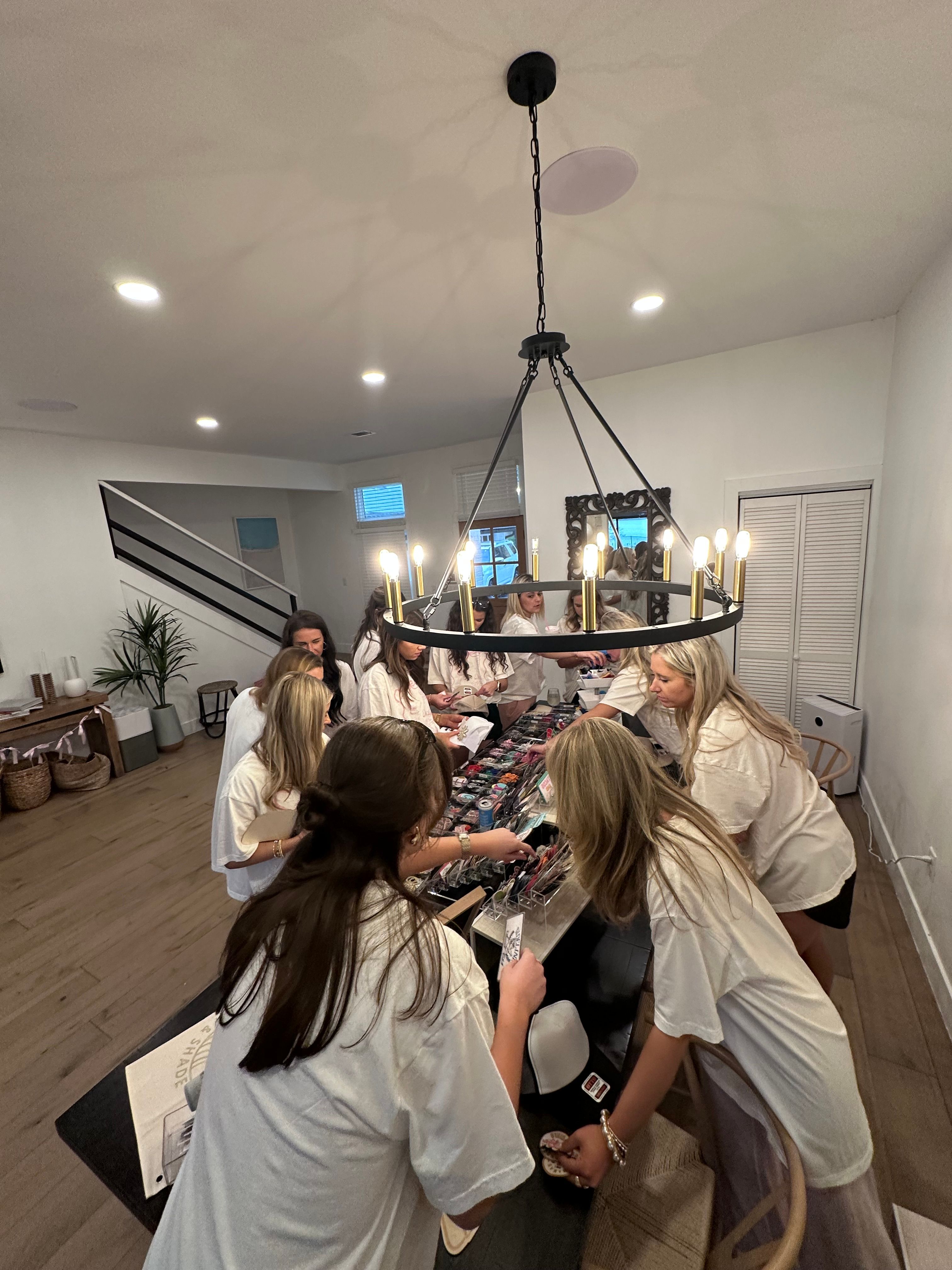 Overhead view of an indoor gathering in a modern home: a group in matching white shirts clustered around a long table picking colorful small items under a black circular chandelier with exposed bulbs.