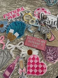 Assorted bridal-themed embroidered and sequin iron-on patches on a weathered wood surface — pink “Future Mrs” and “Put It On My Husband’s Tab” text patches, blue sequin shell, diamond ring, pink gingham heart, daisies, champagne flute and bottle, and disco ball.