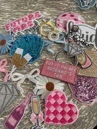 Assorted bridal-themed embroidered and sequin iron-on patches on a weathered wood surface — pink “Future Mrs” and “Put It On My Husband’s Tab” text patches, blue sequin shell, diamond ring, pink gingham heart, daisies, champagne flute and bottle, and disco ball.