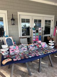 Colorful pop-up market table on a covered patio displaying organized trays of enamel pins, patches, stickers, hair accessories and a cubby rack of pastel caps in front of white French doors and shingled wall.