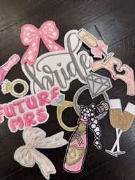 Assorted embroidered bridal patches on dark wood — "Bride" script, "Future Mrs" text, diamond rings, champagne bottle and flutes, pink bows and bachelorette motifs