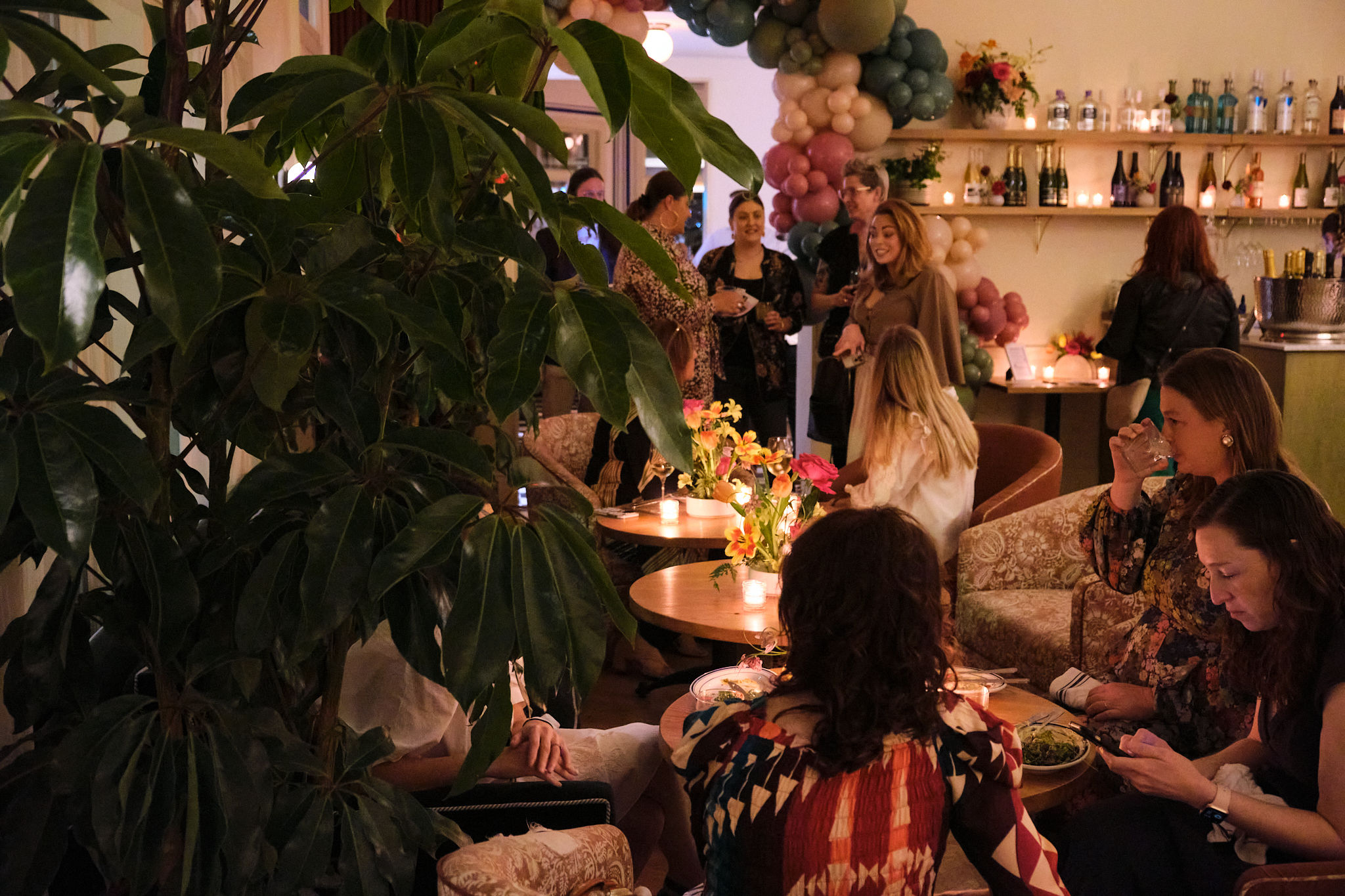 Cozy indoor lounge cocktail party with guests mingling around candlelit tables, floral centerpieces and a pastel balloon arch by a stocked bar, large potted plant in the foreground.