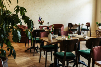Sunlit cozy bistro interior with wooden tables set with glasses and napkins, green velvet chairs, potted plants and small floral centerpieces on parquet floors.