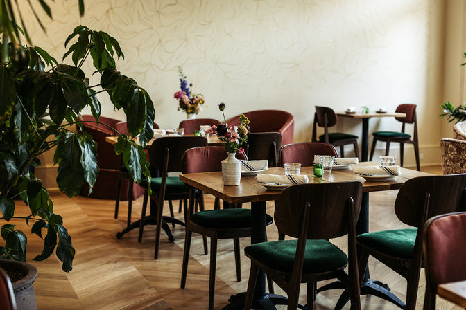 Sunlit cozy bistro interior with wooden tables set with glasses and napkins, green velvet chairs, potted plants and small floral centerpieces on parquet floors.