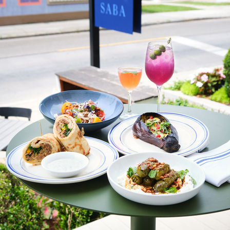 Sunny sidewalk cafe table with a Mediterranean-inspired spread: grilled stuffed eggplant, stuffed grape leaves on yogurt, a savory wrap with dipping sauce, a fresh tomato salad, and two colorful cocktails (purple and peach).