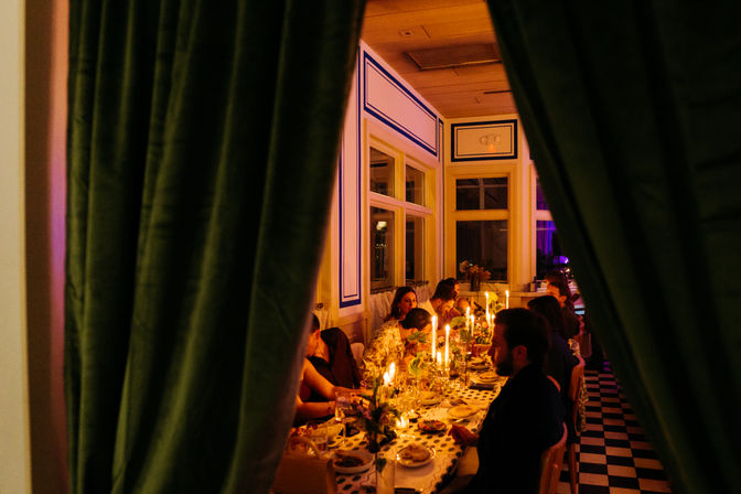 Peek through green velvet curtains at an intimate candlelit dinner around a long banquet table in an elegant indoor dining room with a black-and-white checkered floor.
