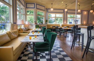 Sunlit cafe interior with green velvet chairs and mustard leather banquette, marble tables on black-and-white checkered tile, large windows and a wood bar.