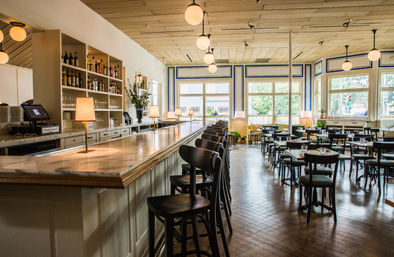 Sunlit restaurant interior with a long marble-topped wooden bar, a row of dark bar stools, globe pendant lights, and a bright dining area of small tables and large windows