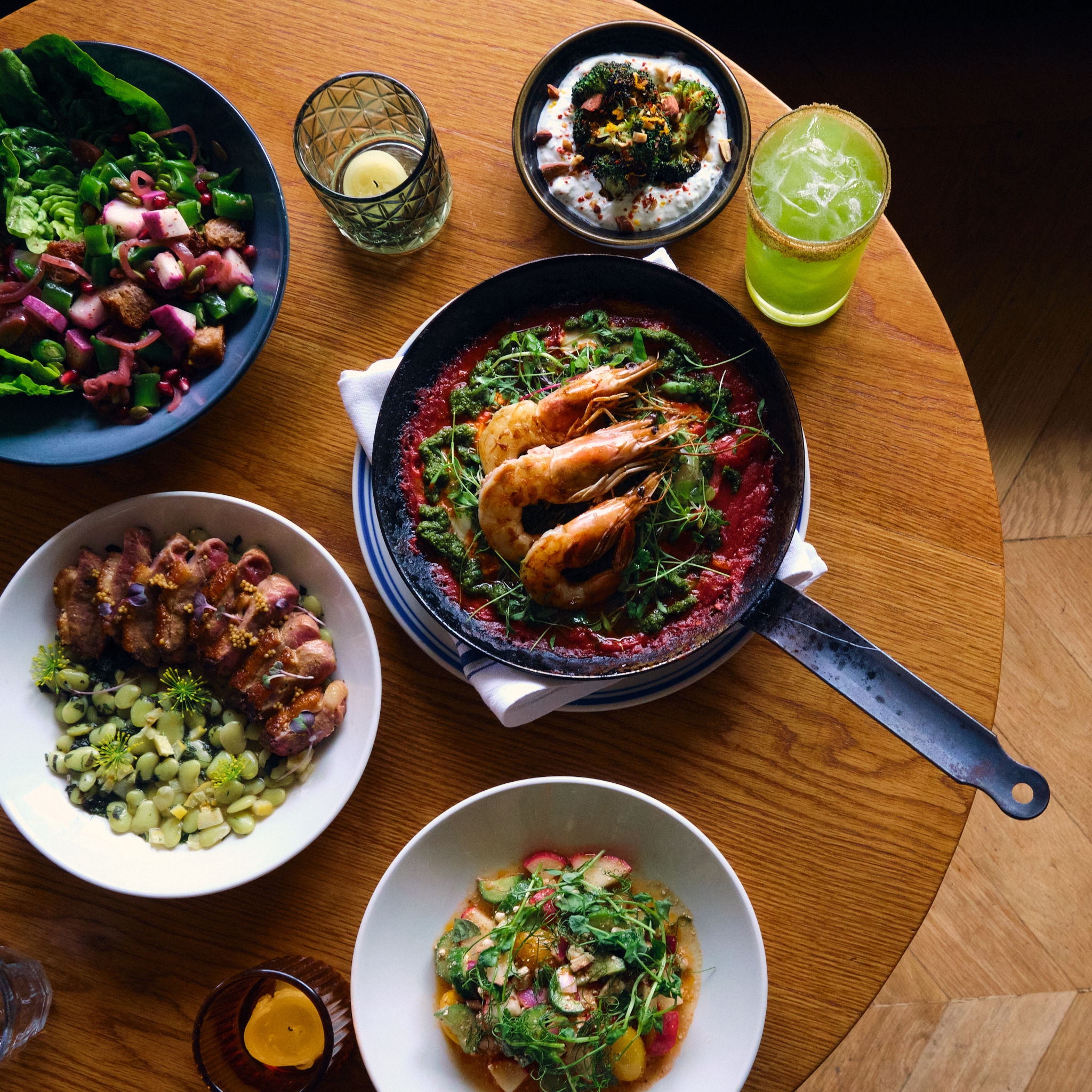 Top-down view of a wooden table with a seafood skillet of prawns in tomato sauce, two fresh salads, a creamy dip and a green cocktail — vibrant bistro-style meal, perfect for seafood lovers.