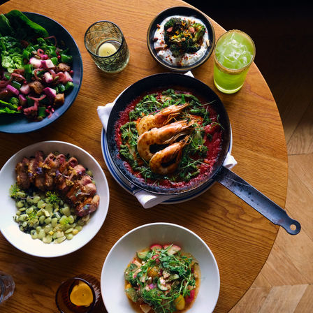 Top-down view of a wooden table with a seafood skillet of prawns in tomato sauce, two fresh salads, a creamy dip and a green cocktail — vibrant bistro-style meal, perfect for seafood lovers.