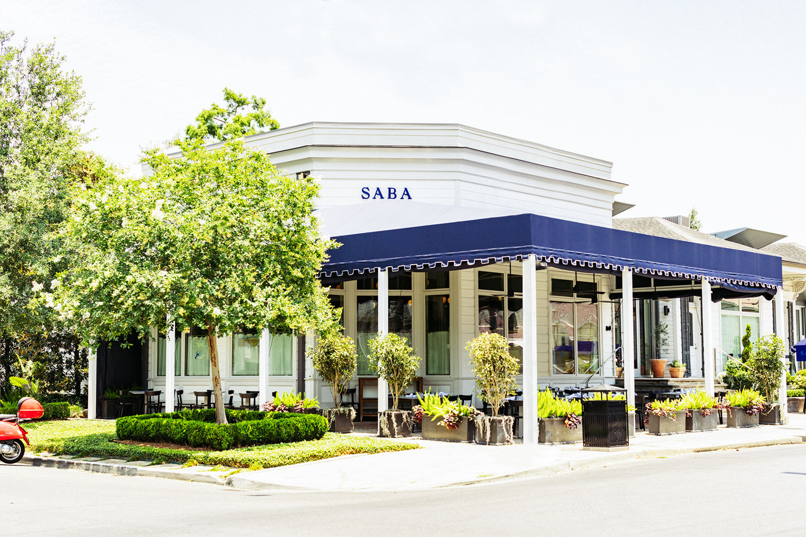 Bright corner café in a white building with a navy awning, outdoor seating surrounded by leafy trees, potted planters, and a red scooter parked on a sunny street.