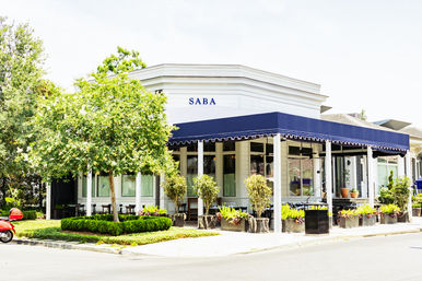Bright corner café in a white building with a navy awning, outdoor seating surrounded by leafy trees, potted planters, and a red scooter parked on a sunny street.