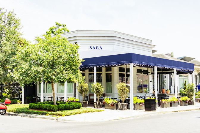 Bright corner café in a white building with a navy awning, outdoor seating surrounded by leafy trees, potted planters, and a red scooter parked on a sunny street.