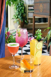 Four colorful craft cocktails—pink spritz, yellow lime highball, coral coupe, and old-fashioned with orange peel—garnished with olives, cherries and herbs on a wooden table in a bright bistro with checkered floor and potted plants