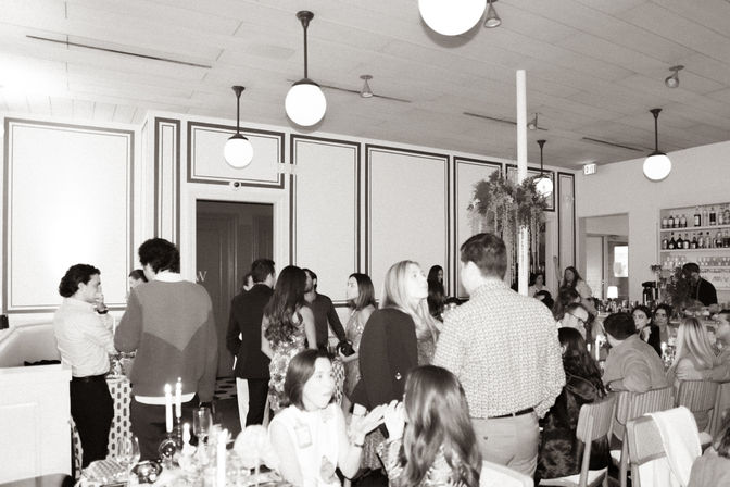 Black-and-white photo of a bustling indoor restaurant event with guests mingling and seated at candlelit tables, bar shelves in the background, globe pendant lights and hanging greenery.