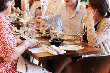 Group of people chatting over white wine and shared small plates at a cozy wooden-table restaurant, casual dinner setting with glasses, plates, and napkins.
