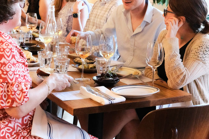 Group of people chatting over white wine and shared small plates at a cozy wooden-table restaurant, casual dinner setting with glasses, plates, and napkins.