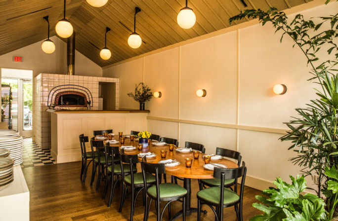 Cozy modern restaurant dining room with a long wooden communal table set for guests, green-cushioned chairs, globe pendant lights, warm wall sconces, tiled wood-fired oven in the back and leafy potted plants.