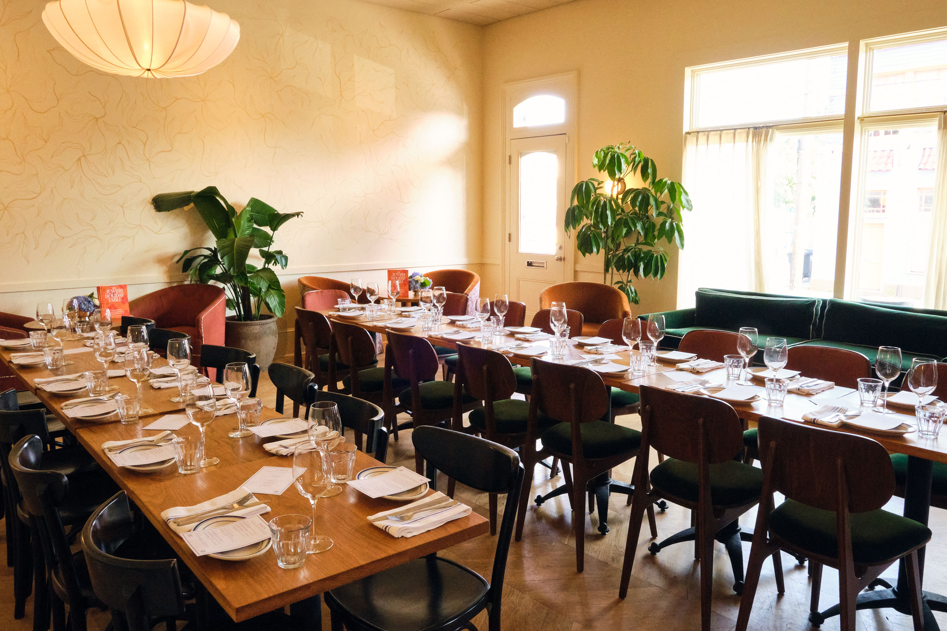 Sunny cozy urban bistro interior with long wooden communal tables set for a group dinner, mid-century chairs, glassware and napkins, potted plants, large windows and a paper pendant light.