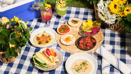 Vibrant outdoor picnic spread on blue-and-white checkered tablecloth with Mediterranean mezze: hummus, olives, assorted dips, pickles, roasted meat slices, fresh vegetable crudités and two floral cocktails surrounded by baskets of sunflowers and mixed blooms.