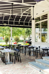 Outdoor dining patio with black chairs and tables, ceiling fans, brick pavers, and a leafy street view