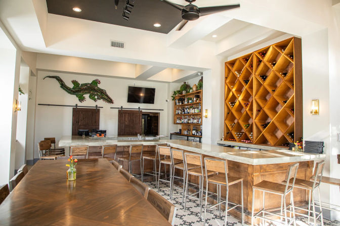 Sunlit modern bar interior with marble-top L-shaped counter and wooden stools, large diamond-pattern wine rack, long communal wood table with small vase, patterned tile floor and mounted alligator wall art.