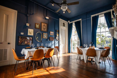 Sunlit restaurant interior with navy-blue walls and tall windows, round tables draped in white linens set with wine glasses, brown leather chairs, hardwood floors, gallery wall and velvet curtains — cozy, stylish dining room.