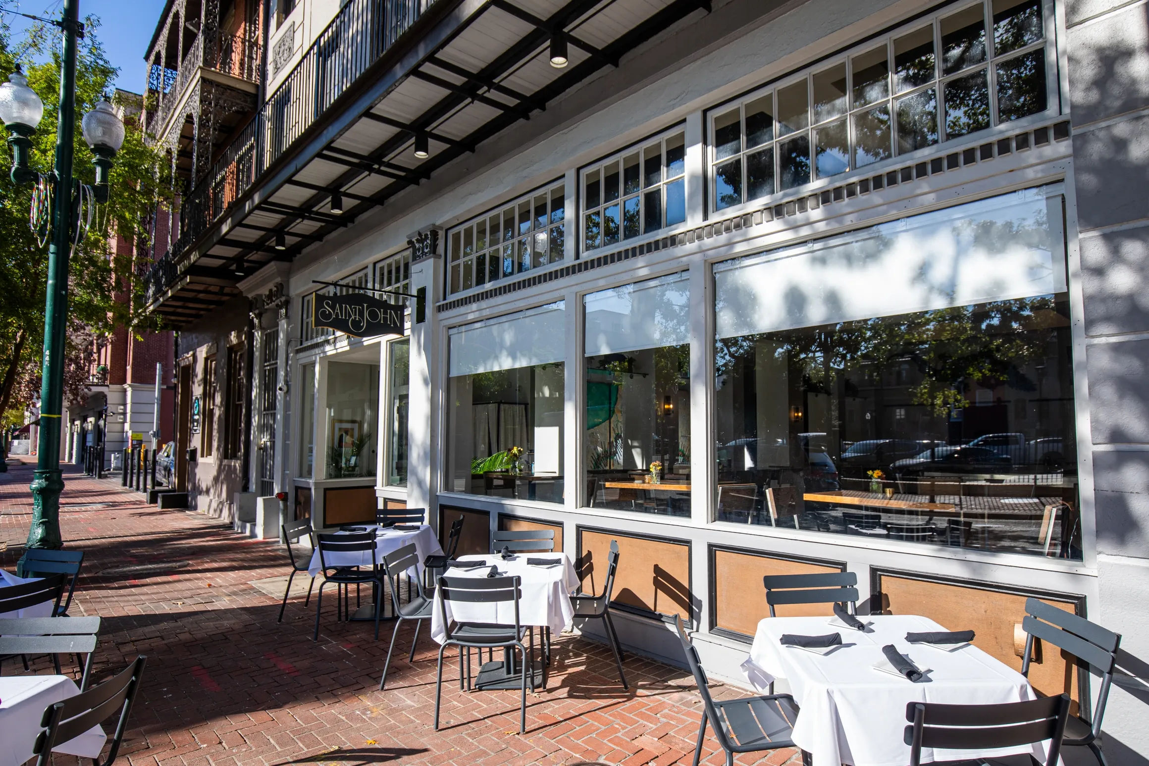 Sun-dappled sidewalk café with white-tablecloth bistro tables and black chairs on a brick-paved downtown street, large storefront windows, iron balconies and vintage street lamps