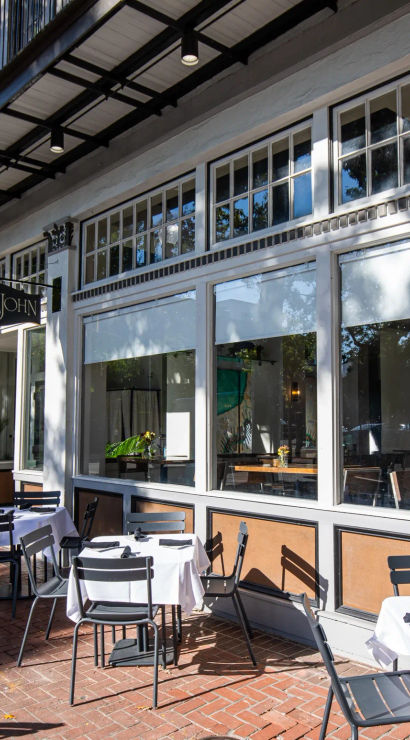 Sun-dappled sidewalk café with white-tablecloth bistro tables and black chairs on a brick-paved downtown street, large storefront windows, iron balconies and vintage street lamps
