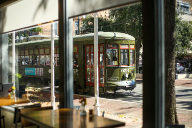 Sunlit vintage green streetcar rolling down a tree-lined downtown street, seen through a café window with empty wooden tables in the foreground.