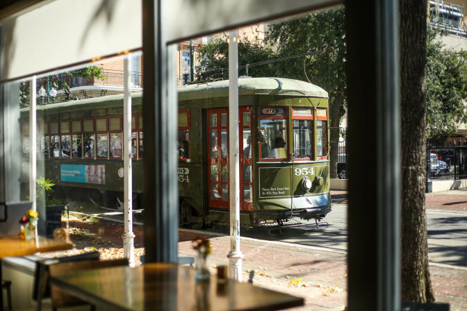 Sunlit vintage green streetcar rolling down a tree-lined downtown street, seen through a café window with empty wooden tables in the foreground.