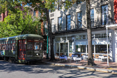 Historic green St. Charles streetcar parked on a sunlit, tree-lined New Orleans street beside a sidewalk café with tables and second-floor balconies casting shadows.