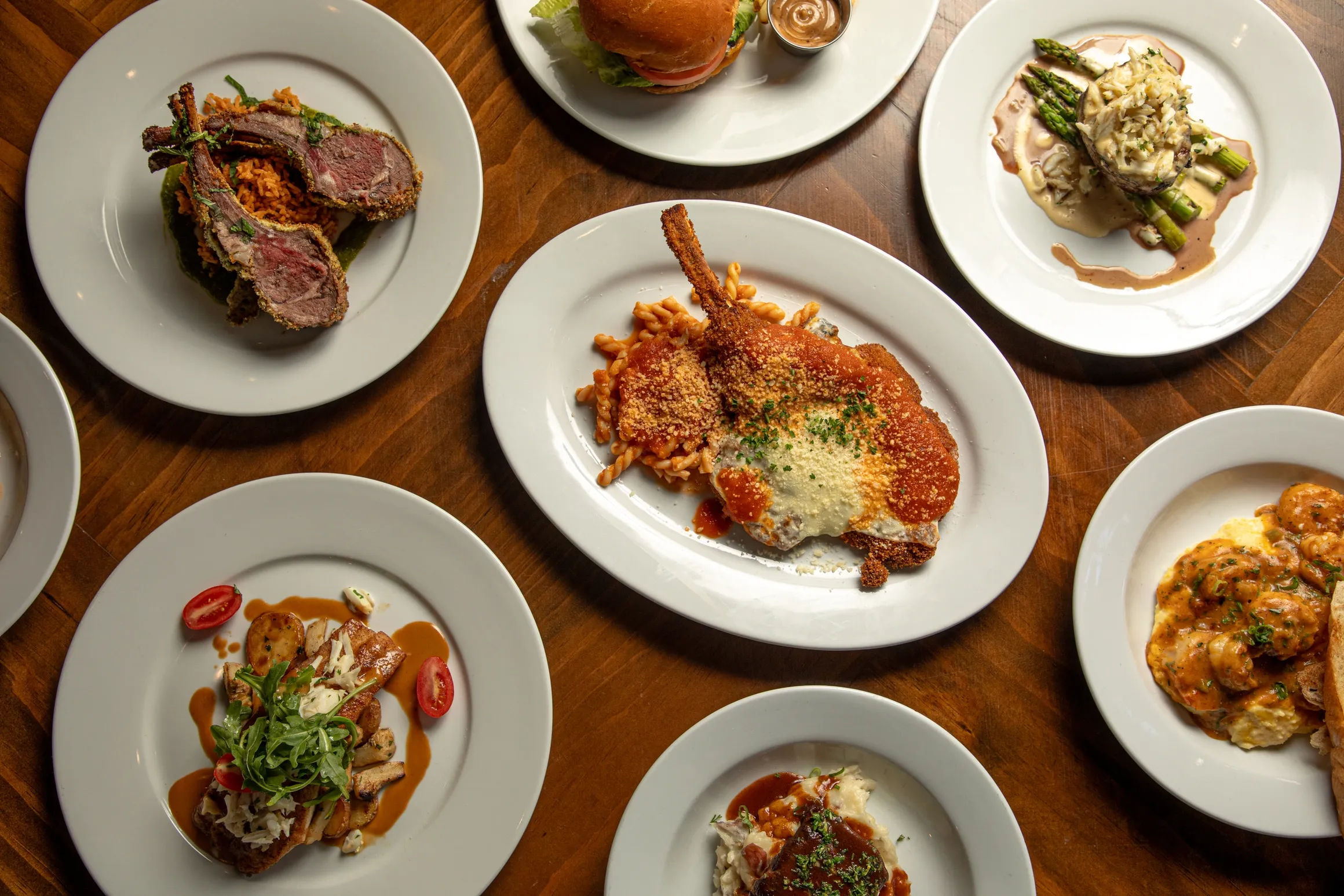 Top-down view of a wooden restaurant table with white plates of assorted gourmet entrees — breaded chop with pasta and red sauce, seared lamb chops, a burger, asparagus with sauce, and creamy shrimp — colorful plated dinner spread.