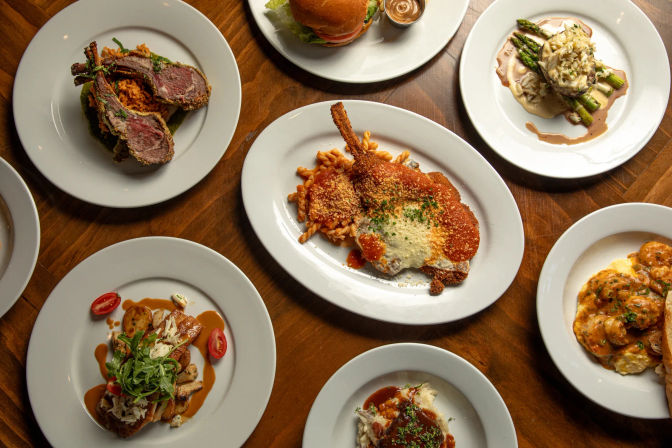 Top-down view of a wooden restaurant table with white plates of assorted gourmet entrees — breaded chop with pasta and red sauce, seared lamb chops, a burger, asparagus with sauce, and creamy shrimp — colorful plated dinner spread.