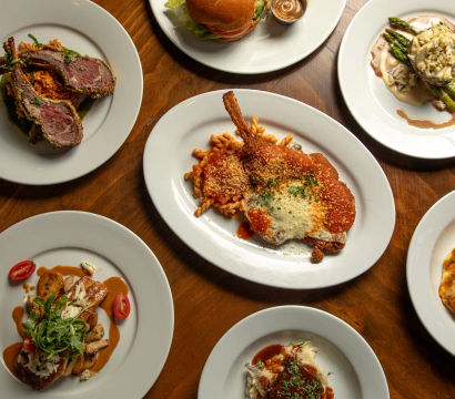 Top-down view of a wooden restaurant table with white plates of assorted gourmet entrees — breaded chop with pasta and red sauce, seared lamb chops, a burger, asparagus with sauce, and creamy shrimp — colorful plated dinner spread.