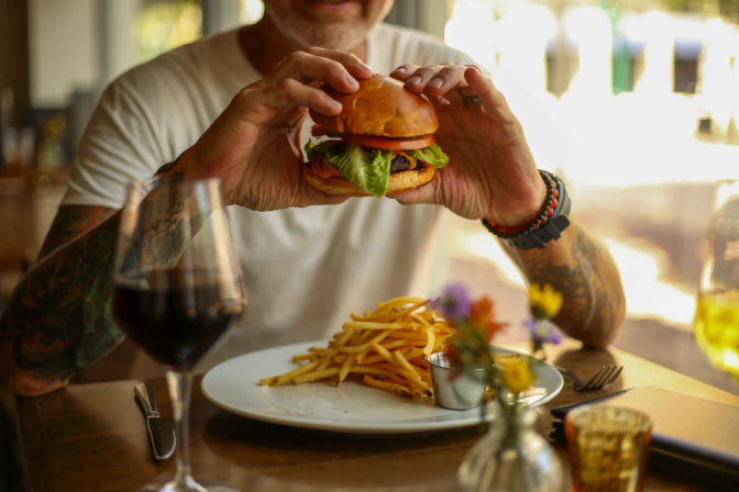 Person with tattooed arms holding a juicy burger with lettuce and tomato above a plate of fries on a casual restaurant table, with a glass of red wine and a small vase of flowers nearby.