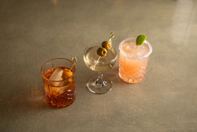 Three cocktails on a concrete bar top: amber glass with a large ice cube and orange twist, a clear coupe garnished with two green olives on a pick, and a pink salted-rim cocktail with a lime wedge.