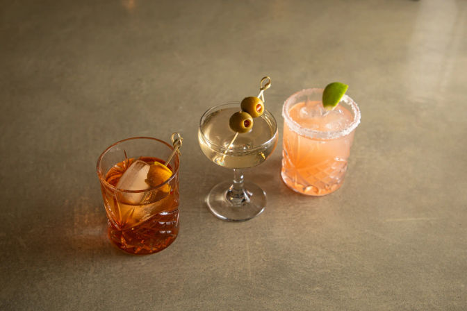 Three cocktails on a concrete bar top: amber glass with a large ice cube and orange twist, a clear coupe garnished with two green olives on a pick, and a pink salted-rim cocktail with a lime wedge.