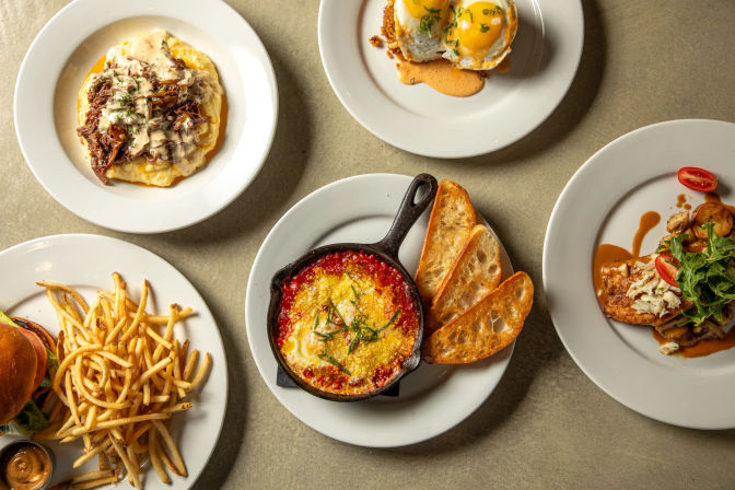 Vibrant brunch spread on a restaurant table: cast-iron skillet of baked eggs in tomato sauce with toasted baguette slices, burger with shoestring fries, two runny eggs over a potato base with hollandaise, shredded meat on creamy grits, and seared chicken with arugula and cherry tomatoes.
