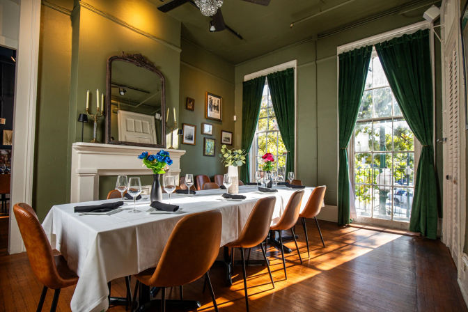 Sunlit private dining room with a long white-tablecloth table set with wine glasses and floral centerpieces, tan leather chairs, green velvet curtains, vintage fireplace and hardwood floors.