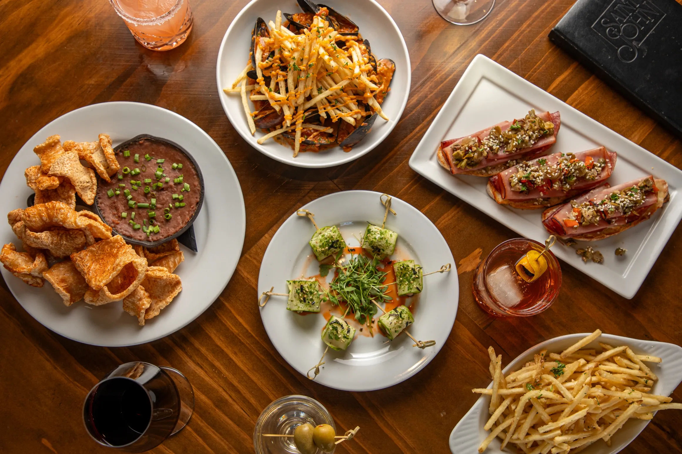 Overhead photo of a wooden table with bistro-style shared plates: crispy pork rinds with black bean dip, mussels topped with shoestring fries, herb-coated cheese skewers, prosciutto-topped crostini, thin French fries, and cocktails and wine — perfect for bar snacks and small-plates dining.