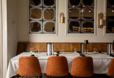 Cozy modern restaurant dining table set with white tablecloth, tan leather chairs, menus and glassware against a wooden banquette and frosted grid mirrors.
